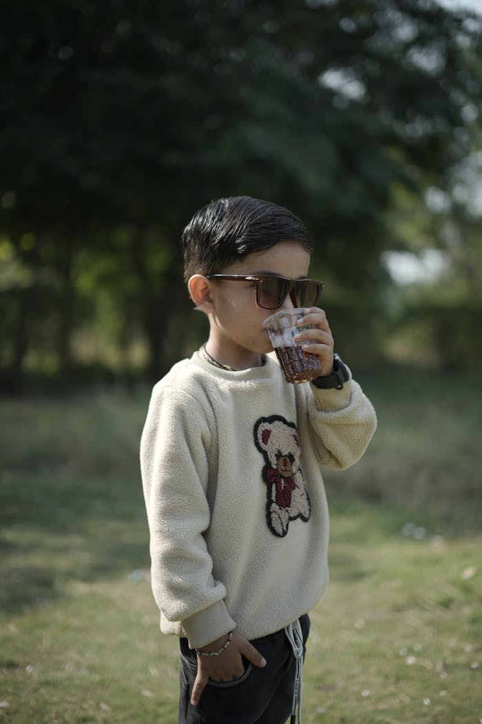 services-01 Young boy with sunglasses sipping a beverage in a park setting, wearing a cozy sweater.